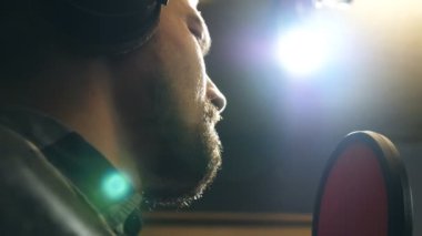 Profile of male singer in headphones singing song into the microphone at sound studio. Young man recording new melody or song. Working of creative musician. Show business concept. Slow motion.