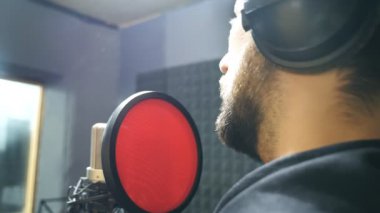 Kulaklıklı sakallı bir erkek şarkıcı ses stüdyosundaki mikrofona şarkı söylüyor. Genç adam yeni bir melodi ya da şarkı kaydediyor. Yaratıcı bir müzisyenin çalışması. Gösteri dünyası konsepti. Yavaş çekim.