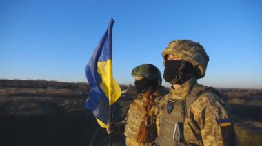 Ukrayna ordusunun genç askerleri ellerinde ulusal bayrağıyla tepenin zirvesinde dikiliyorlar. Kamuflaj üniformalı kadın ve adam Ukrayna bayrağını kaldırıp kırsal alanda gün batımını seyrediyorlar. Kapat..