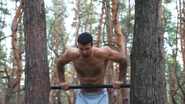 Güçlü ve kaslı bir adam ormanda barfiks çekiyor. Doğada spor yapan cesur bir sporcu. Açık havada duygusal yüz eğitimi almış atletik bir adam. Spor ve aktif yaşam tarzı kavramı.