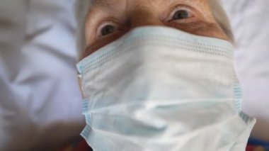 Virüsten koruyucu maske takan yaşlı bir kadının portresi. Hastanede yatan üzgün bakışlı ve tıbbi maskeli hasta kadın. Salgından korunma ya da sağlık hayatı kavramı. Yavaş çekim.