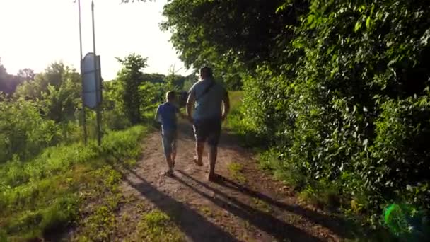 彼の小さな子供と一緒に走る父親は 森の近くの道をスキップしています 彼の小さな息子とパパは 夏の日にトレイルの間を歩いているお互いの腕を持っています パパと子供は一緒にアウトドアで時間を過ごす — ストック動画