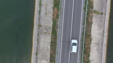 Baraj yolundan geçen beyaz arabanın üst görüntüsü. SUV yaz günü göl kenarındaki yolda gidiyor. Oto üzerinden uçmak nehir ya da gölet köprüsünden geçer. Manzaralı bir yer. Seyahat konsepti. Hava görüntüsü