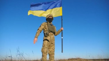 Mavi-sarı pankartıyla mavi gökyüzüne selam veren Ukraynalı erkek asker. Kırsalda Ukrayna bayrağı taşıyan kamuflaj üniformalı genç bir asker. Rus istilasına direniş. Düşük görünüm.