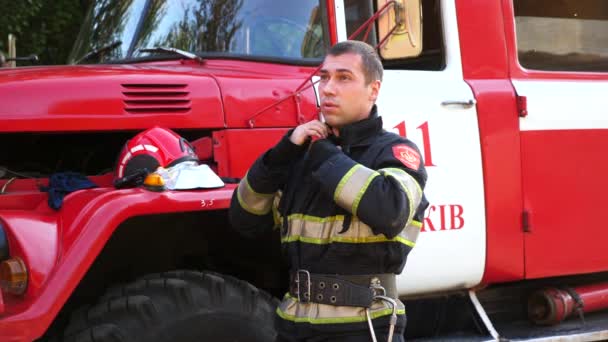 消防車の近くでハードワークした後 若い疲れ消防士は保護ワークウェアを解凍します 大きな赤いトラックを背景に銃撃後の男性消防士 命と英雄的職業を救うという概念 — ストック動画