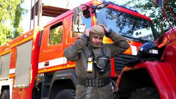 身穿制服脱下巴拉克拉瓦的年轻消防队员站在红色大卡车旁边 在火灾后 男性消防员在消防车的背景下从自己身上取出设备 英雄职业的概念 — 图库视频影像