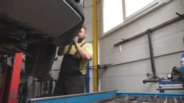 Üniformalı bir tamirci atölyede ya da garajda araba arızalarını tamir ediyor. Araba servisinde kaldırma aracıyla çalışan profesyonel bir tamirci. Düzenli otomobil bakımı kavramı. Yavaş çekim.