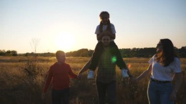 Küçük çocuklu anne ve baba çim tarlasından geçiyorlar. Gün batımında çayırların arasında yürüyen iki çocuklu genç bir çift. Mutlu aile, güzel doğada birlikte vakit geçiriyor. Kapat..