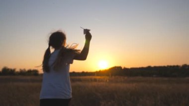Arkadan, elinde kağıt uçakla çim tarlasında koşan şirin bir çocuğun görüntüsü. Mutlu küçük kız çayırların arasında koşuyor ve gün batımında oyuncak uçak fırlatıyor. Çocukluk hayali kavramı.