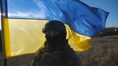 Ukrayna ordusunun kadın askeri doktoru elinde mavi-sarı pankartıyla kameraya bakıyor. Güneşli bir günde Ukrayna bayrağı taşıyan kamuflaj üniformalı ve miğferli bir kızın portresi. Dolly vurdu.