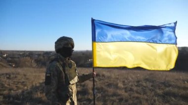 Askeri üniformalı ve miğferinde Ukrayna bayrağı sallayan bir erkek asker resmi. Ukraynalı ordu mensubu ulusal bayrağı kaldırıyor. Rus saldırısına karşı zafer. Yavaş çekim.