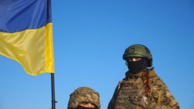 Mavi gökyüzüne karşı çekilen Ukrayna bayrağıyla Ukrayna ordusunun askeri çifti. Kırsalda mavi-sarı pankartlı kamuflaj üniformalı genç askerlerin portresi. İstila direnişi