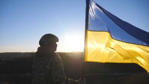 ウクライナ軍の少女は日没を背景に掲揚された国旗で立っている 軍の制服を着た若い女性兵士は 丘のピークにウクライナの手を振ってフラグを保持します 戦争の勝利 ドリーショット — ストック動画