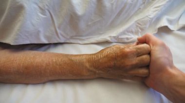Torunun tıbbi klinikteki hasta yetişkin büyükannesinin elini tutması ve dokunması. Genç adam yaşlı büyükannesinin kırışık kolunu hastanede rahatlatıyor. İlgi ve sevgi kavramı. Yavaş çekim.