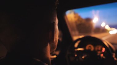 Young man holds hands on steering wheel and rides in auto on highway at night time. Male driver moves in his SUV car on road. Guy drives a modern automobile at evening. Rear view Slow motion.