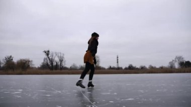 Kız bulutlu bir günde donmuş nehirde ya da gölde paten kayıyor. Doğada buz pateni pistinde kayan artistik paten giyen genç bir kadın. Soğuk kış havasında sporcu kadın eğitimi. Kış zamanı aktif bir eğlence. Yavaş çekim.