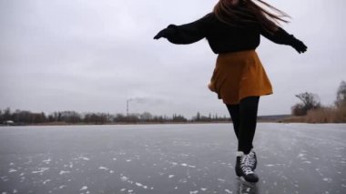 Bulutlu bir günde donmuş nehirde kayan sporcu kadın. Doğada buz yüzeyinde kayan buz pateni giymiş bir kadın. Genç kız soğuk kış havasında gölde aktif boş vakit geçiriyor. Yavaş çekim.