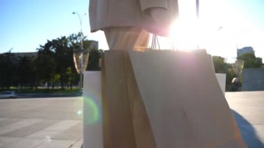 Hands of young woman carry shopping bags walking along urban street. Female arms hold paper packages going on city square after purchases. Concept of sales and discounts. Slow motion Close up.