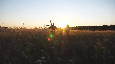 Young parents with little cute girl holds hands of each other running among summer meadow at sunset. Mother and father with small daughter jogging with multicolored kite through grass field. Close up.