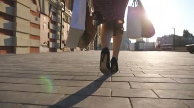 Paketlerle şehir meydanına giden bir bayan. Şehir caddesinde yürüyen genç kadınların kolları alışveriş çantaları taşıyor. Siyah ayakkabılı, yüksek topuklu bir kadın kaldırıma çıkıyor. Arka görünüm.