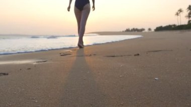Gün batımında okyanus kıyısında kumsalda yürüyen mayo giymiş genç bir kadın. Güzel genç kız dalgalarla deniz kıyısına gidiyor ve tatilin tadını çıkarıyor. Yaz tatilinde dinlenme kavramı. Yavaş çekim.