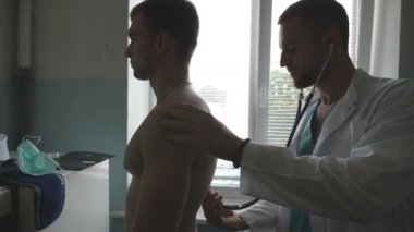 Doktor, hastanedeki ofisteki adamın arkasını kontrol ediyor. Atletik adamın kalp atışlarını dinleyen bir tıp çalışanı. Doktor steteskoplu genç bir erkek hastayı muayene ediyor. Kapat..