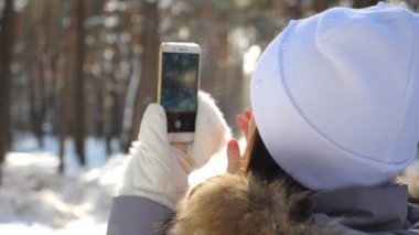 Unrecognizable girl in white cap taking photo of scenic snowy forest on her smartphone. Woman using phone to get beautiful pictures of winter landscape. Lady enjoying holiday at wintertime. Slow mo.