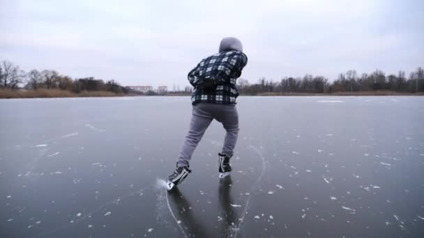 曇った日に凍った川でスケートをする若者 フィギュアスケートの男性スケーターは 屋外の氷表面にスライディングします 冬の寒い冬の湖でのスポーツマントレーニング 冬季のアクティブレジャー — ストック動画