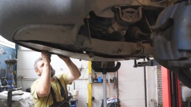 Sakallı tamirci atölyeye özel bir aletle motor koruması kuruyor. Profesyonel oto tamircisi garajdaki bir kaldırma aracının altında çalışıyor. Otomobil bakımı kavramı. Yavaş çekim