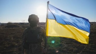 Ukrayna ordusunun askeri sahada mavi-sarı bayrağı kaldırıyor. Kamuflaj üniformalı erkek asker Ukrayna bayrağı sallıyor. Rus saldırısına karşı zafer. İstila direnişi.