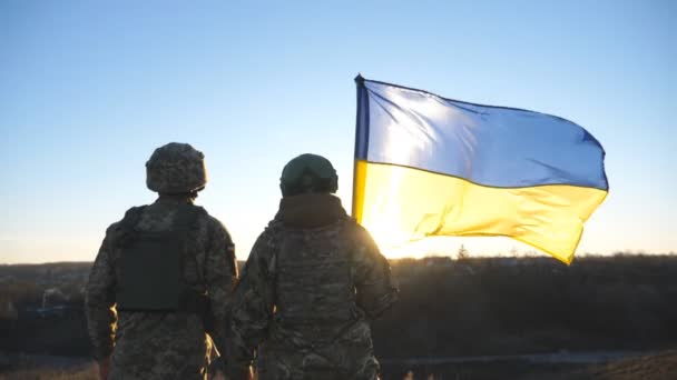 ウクライナ軍の軍のカップルは 手を振ってウクライナの旗で丘のピーク時に立ってお互いの手を保持します 迷彩服の若い兵士たちは日没に対する国旗を掲揚した — ストック動画
