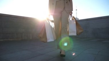 Attractive woman holds shopping bags going at city street after purchases. Beautiful young girl carries paper packets walking along town square. Concept of seasonal sales and discounts. Slow motion.