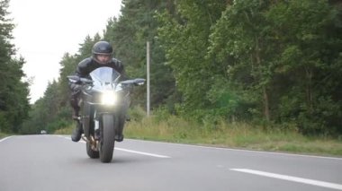 Orman yakınlarındaki kırsal yolda, modern spor motosikletli bir adam. Motosikletli, kır yolunda motosikletiyle yarışıyor. Yolculuk sırasında bisiklet süren adam. Özgürlük yaşam tarzı ve macera kavramı. Yavaş çekim.