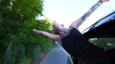 Araba camından sarkan dövmeli neşeli punk kızı ve yolculuk boyunca eğleniyor. Pembe saçlı mutlu hippi kadın şehir yolunda araba sürerken eğleniyor. Seyahat kavramı.