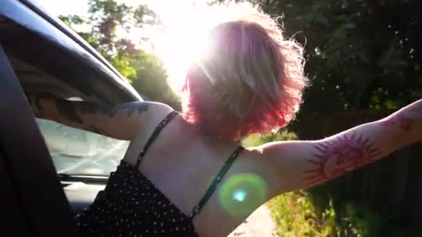 タトゥーを持った若いパンクガールは 自動車の窓から漏れ出し 国道を走りながら手を握ります 田舎で車で運転しながらピンクの髪を楽しんでいるヒッピー女性 — ストック動画