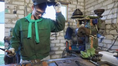 Erkek kaynakçı koruyucu maske takıyor ve atölyede çalışmak için maskesini indiriyor. Eldivenli tamirci lehimleme demiri alıyor ve metal parçaları garajda kaynak yapmaya hazırlanıyor. Dolly vuruldu. Yaklaş..