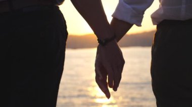 Gün batımında erkek ve kadın ellerinin silueti bir deniz arka planına karşı birbirlerini tutuyorlar. Dışarıda kollarını birleştiren genç bir çift. Sevgi ve mutluluk kavramı. Arka plan görünümünü kapat Yavaş çekim.