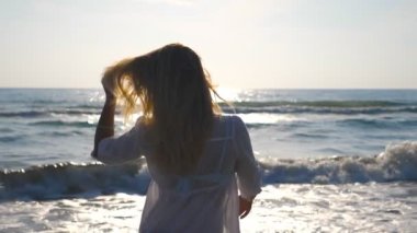 Bikini ve tişörtlü genç bir kızın profili kıyıda yürüyor ve sarı saçlarıyla oynuyor. Yaz tatilinde sahilde eğlenen güzel bir kadın. Arka planda deniz manzarası. Yavaşça kapat.