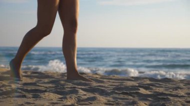 Güneşli bir günde sahilde çıplak ayakla yürüyen bir kadın. Genç kız bacakları deniz sahilinde geziyor. Arka planda deniz manzarası. Yaz tatili ya da tatil konsepti. Yavaşça kapat..
