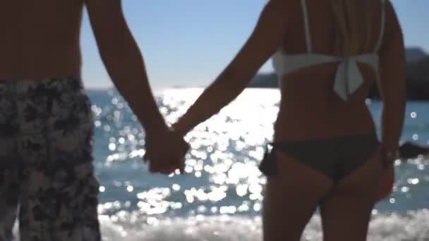 美丽的一对手牵着手 在阳光灿烂的日子走向大海 年轻夫妇在夏天的旅行中在一起 背景上水面上的阳光反射 靠近点 — 图库视频影像