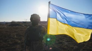Ukrayna ordusunun askeri sahada mavi-sarı bayrağı kaldırıyor. Kamuflaj üniformalı erkek asker Ukrayna bayrağı sallıyor. Rus saldırısına karşı zafer. İstila direnişi.