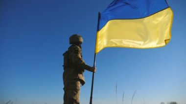Kamuflaj üniformalı bir adam mavi gökyüzüne karşı Ukrayna bayrağı sallıyor. Ukrayna ordusunun erkek askeri ulusal bayrağı kaldırıyor. Rus saldırganlığına karşı zafer. Düşük görünüm.
