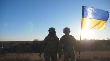Askeri üniformalı genç kadın ve adam gün batımında çayırda mavi-sarı pankartıyla gidiyorlar. Ukrayna bayrağıyla yürüyen Ukrayna ordusunun kadın ve erkek askerleri. Rus işgaline direniş