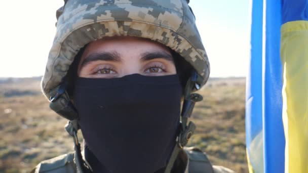 用头盔和巴拉克拉瓦紧盯着镜头的男兵棕色的眼睛 在农村看到一个年轻的军人拿着蓝色和黄色的旗帜 乌克兰入侵领土的概念 — 图库视频影像
