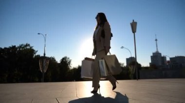 Elegant young lady holds shopping bags going at city square after purchases. Attractive woman carries paper packets walking along sunny urban street. Sales and discounts concept. Slow motion.