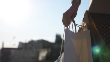 Female arm holds full paper packets walking along urban street. Hand of young woman carries shopping bags going on city alley after purchases. Concept of seasonal sales and discounts. Close up Slow mo