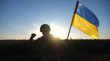 Soldier of ukrainian army stands at hill and holds waving flag of Ukraine. Man in military uniform and helmet lifted up flag against sunset. Victory against russian aggression. Invasion resistance.