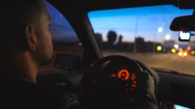 Rear view of young man holds hands on steering wheel and drives a modern automobile at evening. Male driver moves in his SUV car on road. Guy rides in auto on highway at night time. Close up Slow mo.