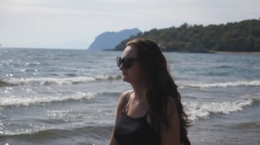 Dişi gezgin, okyanus kıyısında güzel manzaralara hayran gezen güneş gözlükleridir. Manzaralı deniz manzarasının ve tatil beldesinin tadını çıkaran genç bir kız. Yaz seyahati kavramı. Yavaş çekim. Yakın çekim..