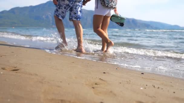 一对年轻夫妇的腿在大海背景下沿着海滩站在一起 在海边散步 泼水洗脚 男人和女人的腿在沙滩上波涛汹涌 靠近点 — 图库视频影像